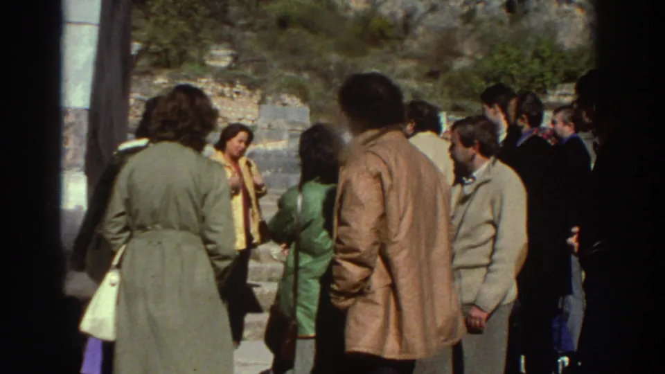 8mm frame showing a larger portion of the visitor group at ancient Greek ruins, stone wall and tree-lined hillside in background, c.1976.