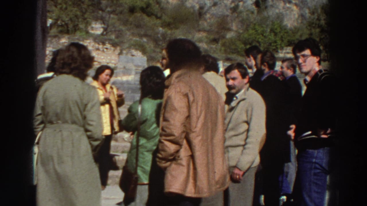 Group of adults in 1970s trench coats and casual wear gathered at an outdoor archaeological site, hills visible behind.