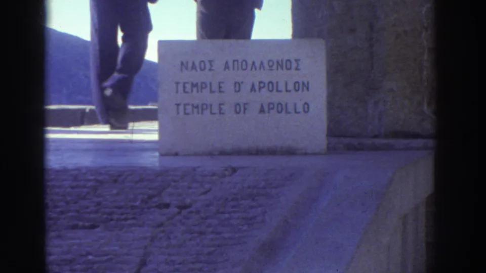 Low-angle shot of the trilingual stone site marker at the Temple of Apollo; visitor's legs pass at left edge, Greece, 1976.