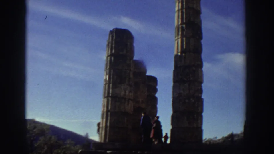 Remaining columns of the Temple of Apollo against a clear blue sky; small visitor figures at the base, mountains visible behind, Greece, 1976.