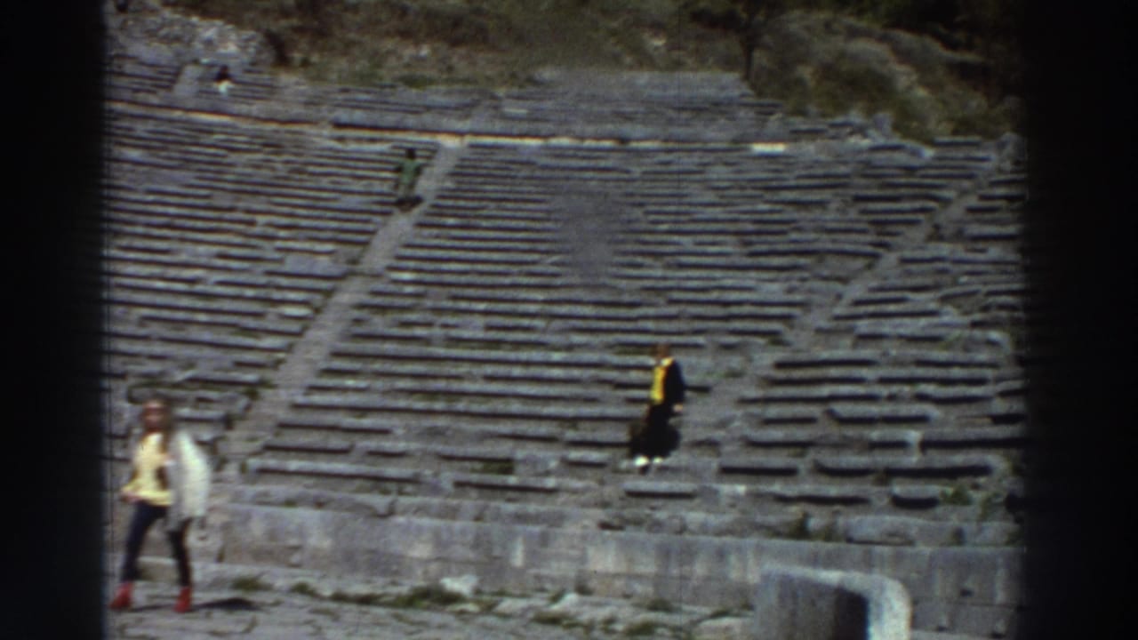 Two tourists on the worn stone seating rows of an ancient Greek amphitheater, circa 1976; one in a yellow jacket stands out against grey stone.