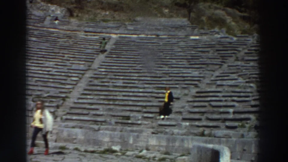 Two tourists on the worn stone seating rows of an ancient Greek amphitheater, circa 1976; one in a yellow jacket stands out against grey stone.