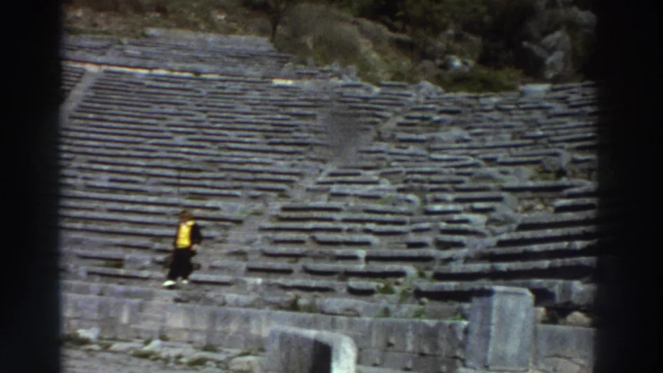 A tourist in a yellow jacket ascends tiered stone seating of an ancient Greek amphitheater, captured on 8mm film circa 1976.
