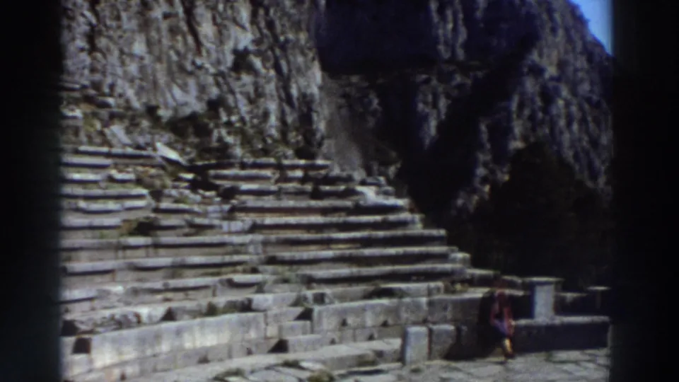 Lower stone steps and orchestra area of an ancient Greek theater with a dramatic sheer limestone cliff backdrop and blue sky; a tourist in red stands at the base, circa 1976.