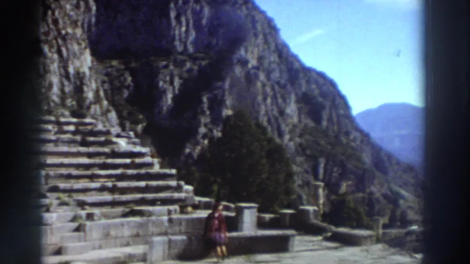 Lower seating rows and orchestra steps of an ancient Greek theater, framed against a sheer limestone cliff and blue sky; a tourist in red looks upward, circa 1976 8mm.