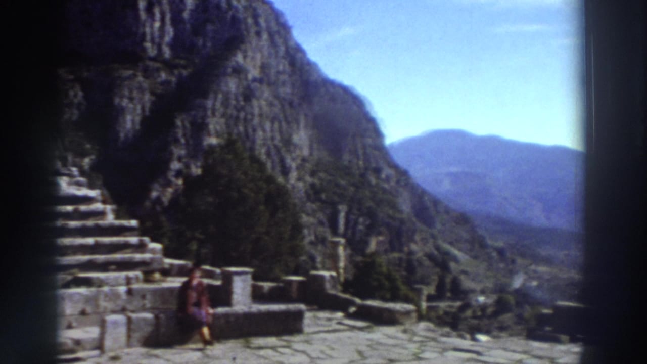 Visitor in bright 1970s clothing rests on a stone terrace at a Greek archaeological site, dramatic rocky cliffs and a deep valley beyond; Super 8, Greece, 1976.