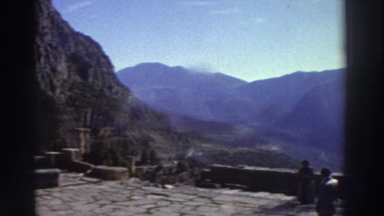 Wide stone-paved terrace at ancient ruins with a figure at far right, steep cliffs and a deep mountain valley in the background.