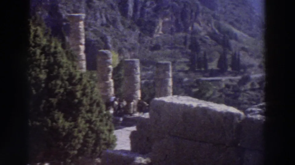 Truncated column drums and large stone blocks at a Greek archaeological site, 1976, with dark cypress trees and visitors visible at the base of the ruins.