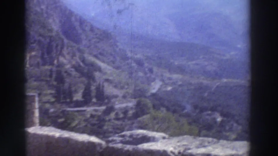 Panoramic view of a deep Greek mountain valley from an ancient ruins plateau, 1976, stone ruin walls and cypress trees in the foreground, blue-hazy peaks beyond.
