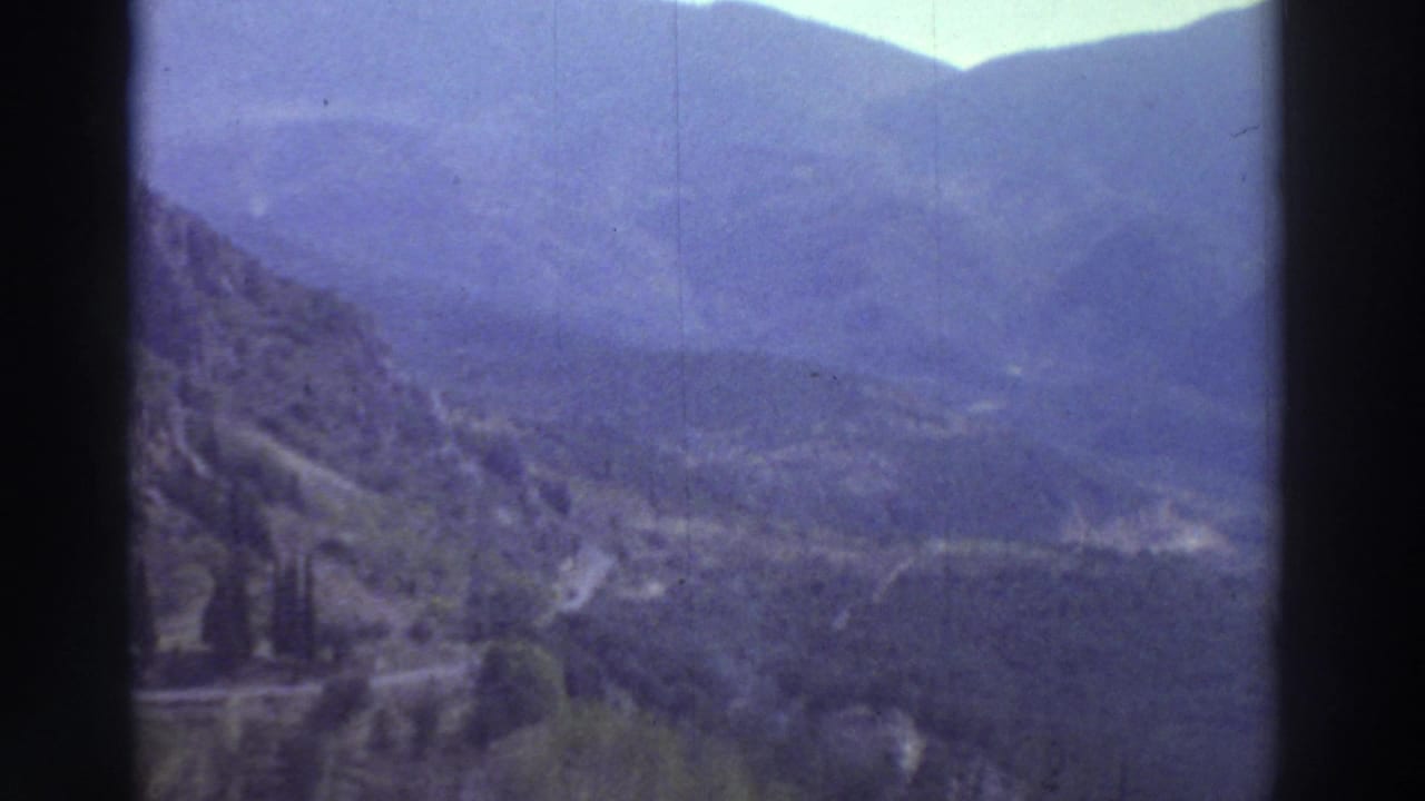 8mm home movie frame from Greece, circa 1976, showing sweeping mountain panorama with pronounced blue-purple color cast from film dye aging.