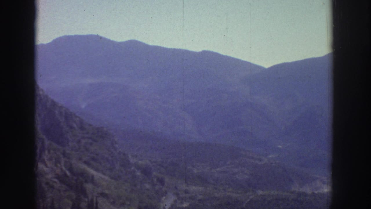Wide shot of steep mountain ridges with deep blue haze; a faint winding road visible in the valley below.