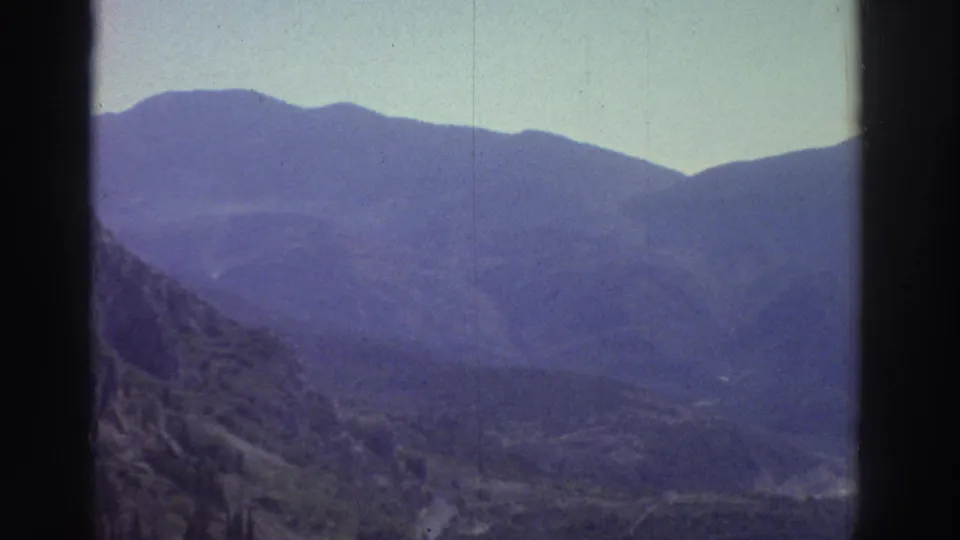Continued 8mm pan across Greek mountain terrain, circa 1976, with a narrow road threading through the valley floor and strong purple-blue film fading across the frame.