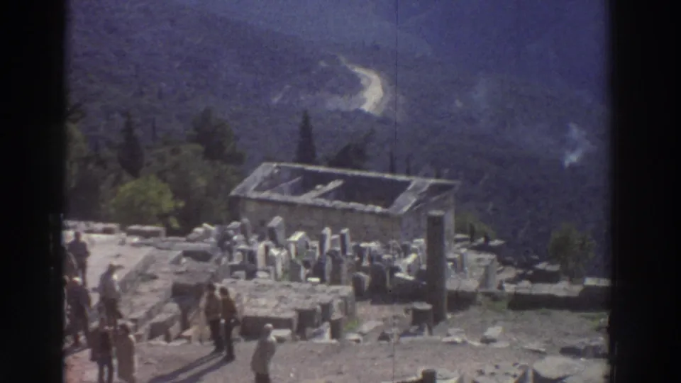 8mm home movie frame from a Greek archaeological site, circa 1976, showing scattered stone blocks, column drum bases, a metal conservation shelter, and tourists amid dramatic mountain scenery.