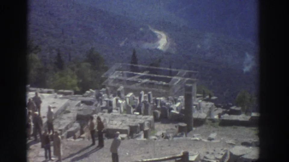 8mm frame of a Greek archaeological site, circa 1976, with tourists walking among scattered stone blocks and column stumps; a protective metal structure is visible over part of the ruins with dramatic mountain backdrop.