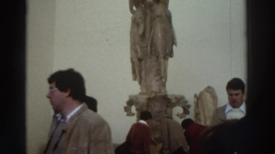 1970s Greece — museum gallery interior with classical marble statue on pedestal; male tourist in brown jacket and other visitors in foreground.