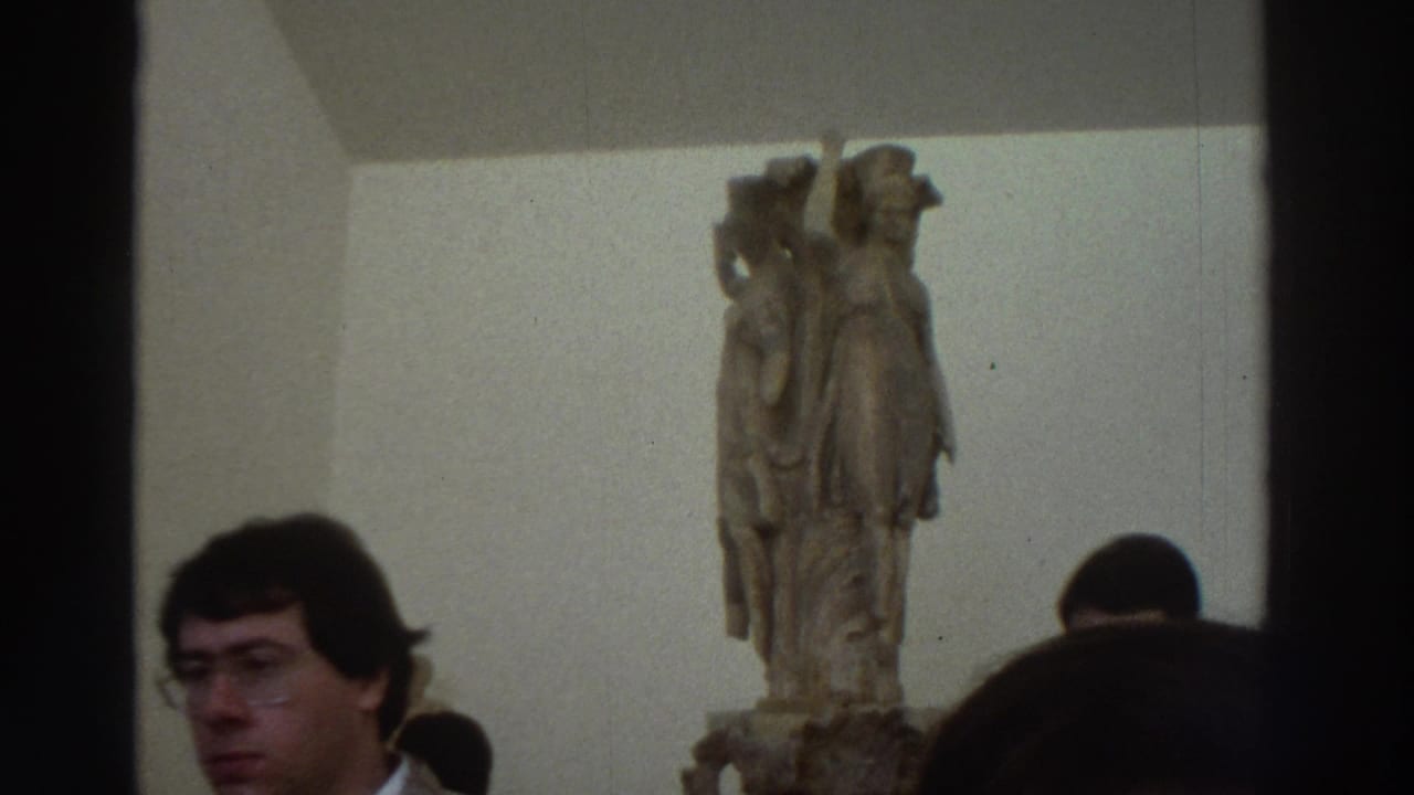 Man with glasses and dark hair close to camera in museum gallery; classical marble statue visible behind him.