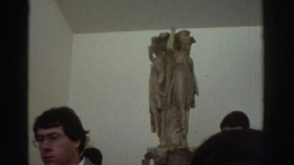 1970s Greece — close view of male museum visitor with glasses; large robed classical marble statue on pedestal in background.