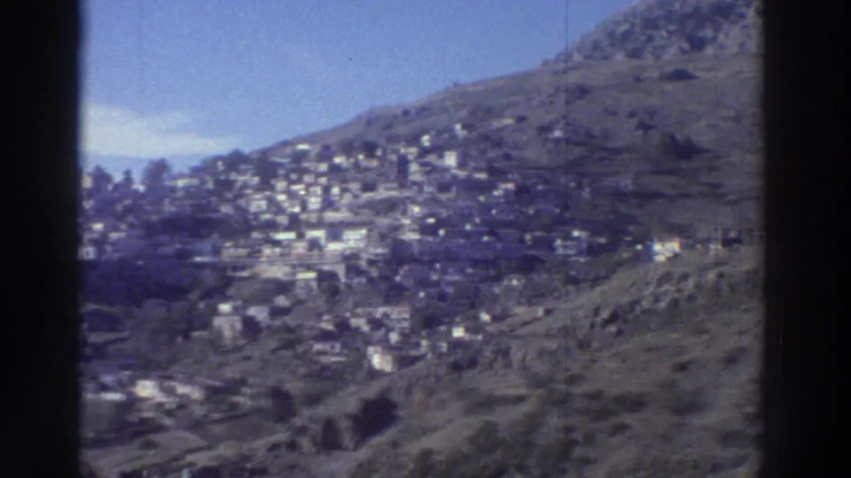 Greece, circa 1976 — hillside settlement with white buildings spread across rocky slopes, blue sky above; shot on 8mm with slight blue-purple color cast.