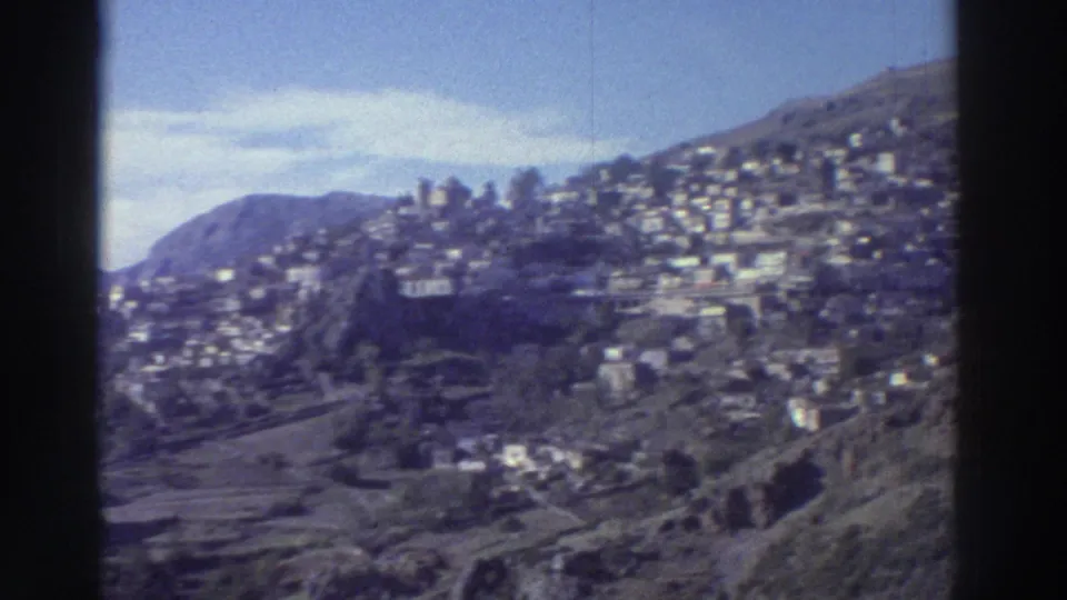 Greece, circa 1976 — panoramic view of white buildings on rocky hillside with blue sky and partial cloud cover; 8mm film with characteristic color rendering.