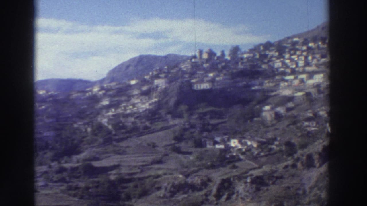 Whitewashed village houses scattered across a rocky terraced hillside under a pale blue sky, filmed on Super 8.