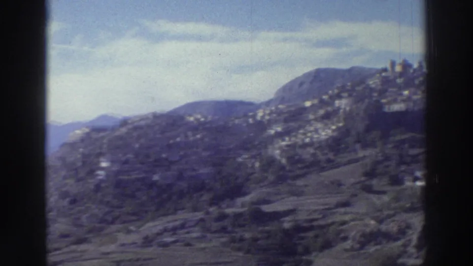 Mid-pan view of a Greek mountain village on a rocky hillside, circa 1976; whitewashed buildings on terraced slopes under a hazy sky.
