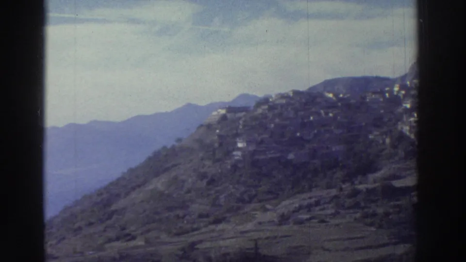 Panoramic view of a Greek hillside village on a rocky summit, circa 1976, with layered mountain ridges receding into a hazy blue valley.