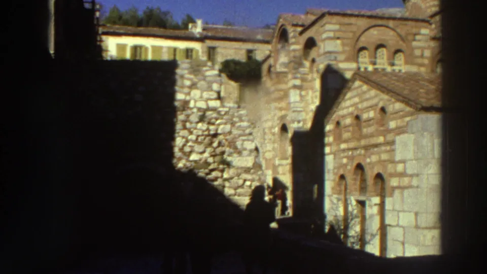 Stone village with Romanesque-arched windows and silhouetted visitors in foreground, filmed on 8mm in Greece, circa 1976.