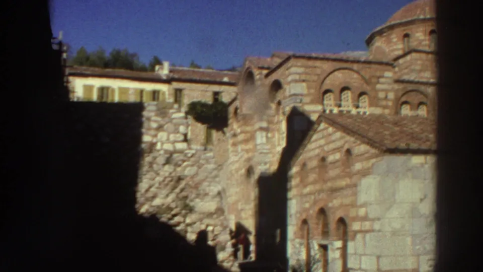 Opening frame of 8mm tourist footage of a Byzantine-style church in Greece, 1976; stone masonry facade, terracotta dome, and a visitor figure near the doorway