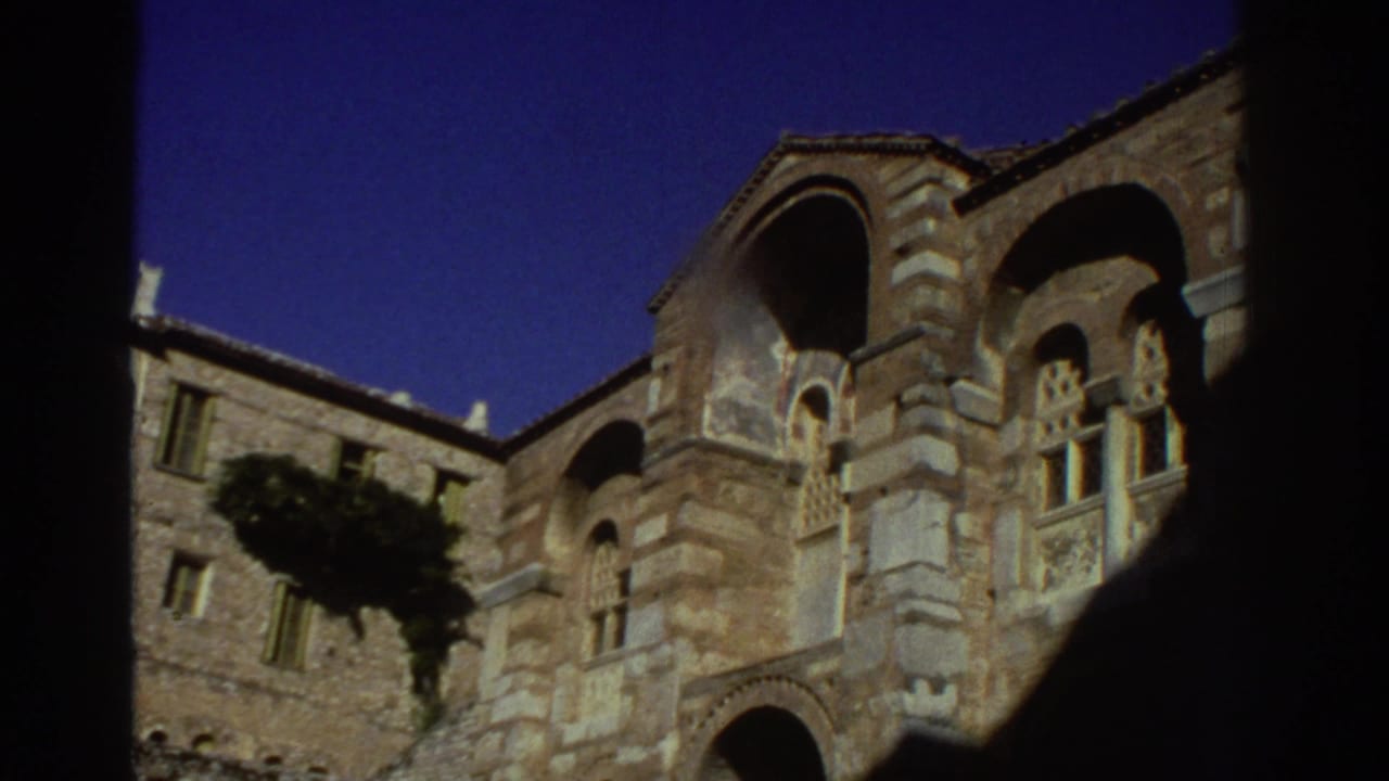 Upward view of a multi-story Byzantine stone church exterior in Greece, circa 1976, framed by a dark foreground silhouette; shot on 8mm film.