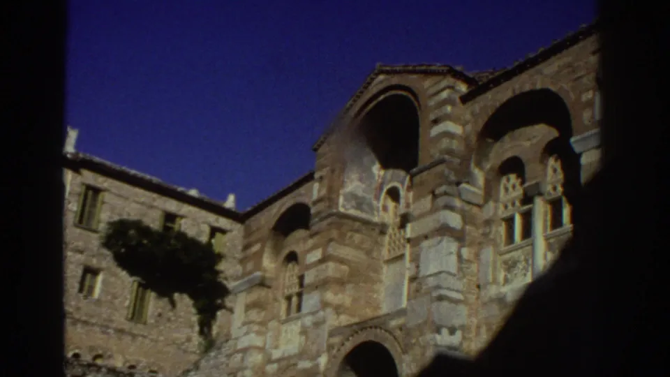 Upward view of a multi-story Byzantine stone church exterior in Greece, circa 1976, framed by a dark foreground silhouette; shot on 8mm film.