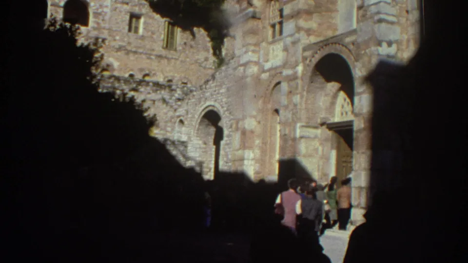 Several visitors, including one in a light-colored top, gather near the Byzantine church entrance in Greece, circa 1976, captured on 8mm film.