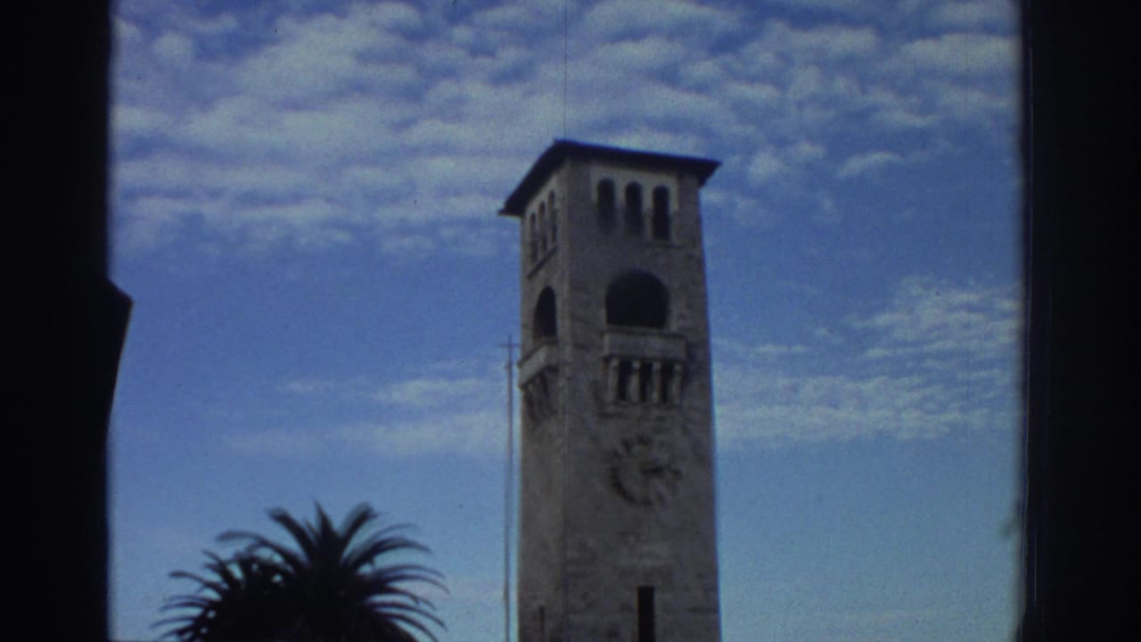 Multi-story stone clock tower in Greece, circa 1976, seen from below against a blue sky; palm tree at frame left; 8mm home-movie grain visible.