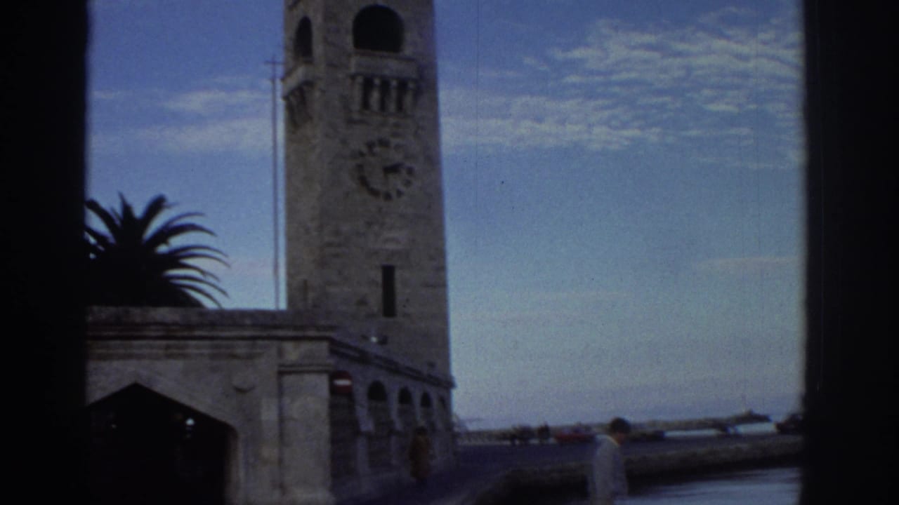 Stone clock tower and arcaded stone base building at a Greek harbor circa 1976, with clock face, palm tree, and a red vehicle visible at lower right.