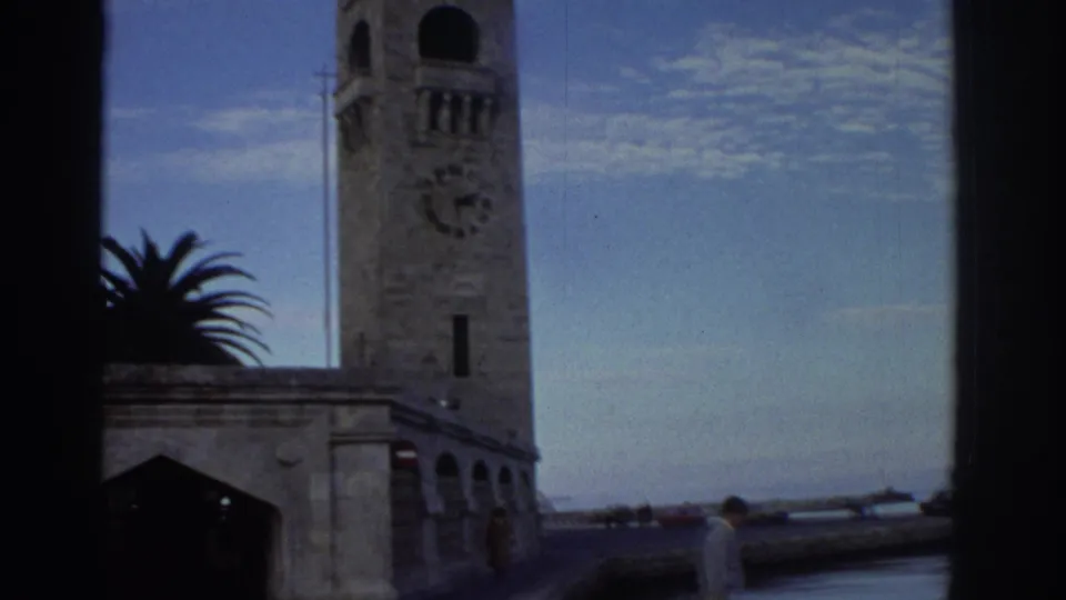 Stone clock tower and arcaded stone base building at a Greek harbor circa 1976, with clock face, palm tree, and a red vehicle visible at lower right.