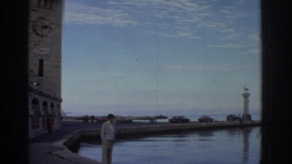 Greek coastal port quay circa 1976: lone adult pedestrian, parked 1970s cars, calm harbor water, and lighthouse pillars at the breakwater; tower partially visible at left.