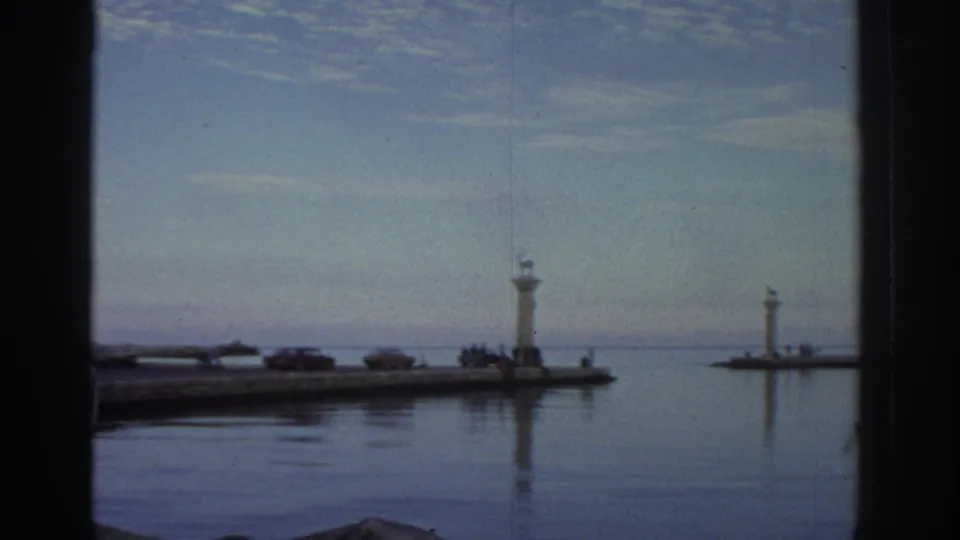 Greek port harbor entrance circa 1976 showing two lighthouse columns with small animal figures on top, calm flat water, and a pier extending to sea.