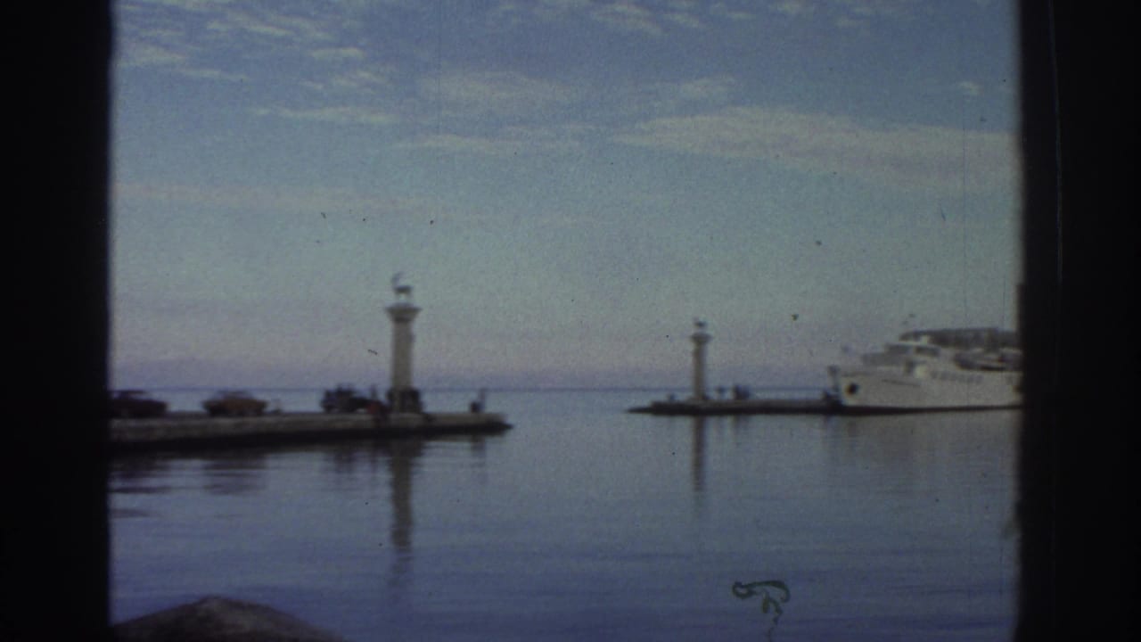 8mm frame from a 1976 Greece trip showing a calm harbor through a window, with twin pier lighthouse markers and a white ferry at anchor.