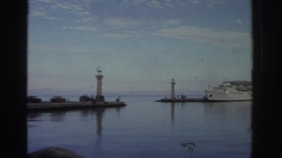 8mm frame from a 1976 Greece trip showing a calm harbor through a window, with twin pier lighthouse markers and a white ferry at anchor.
