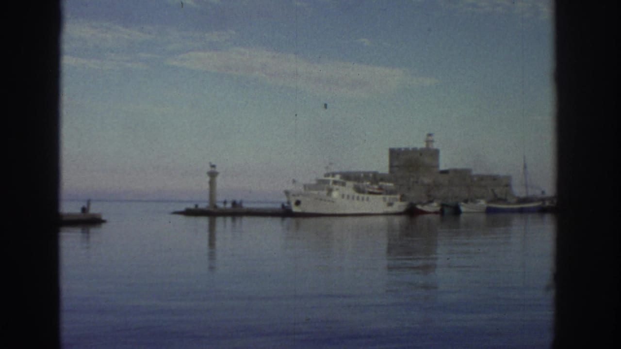 Camera pan reveals a stone fortress at the harbor's right edge, with twin lighthouses and white ferry still visible at center.
