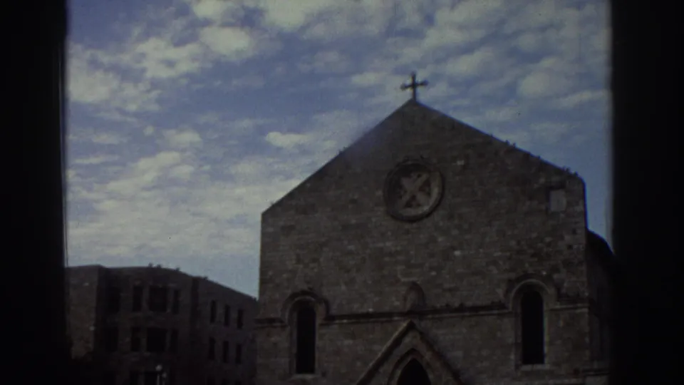 8mm frame from 1976 Greece showing a stone Gothic-style church with gabled facade, circular rose window, cross at peak, and Gothic arched windows, viewed through a window frame.