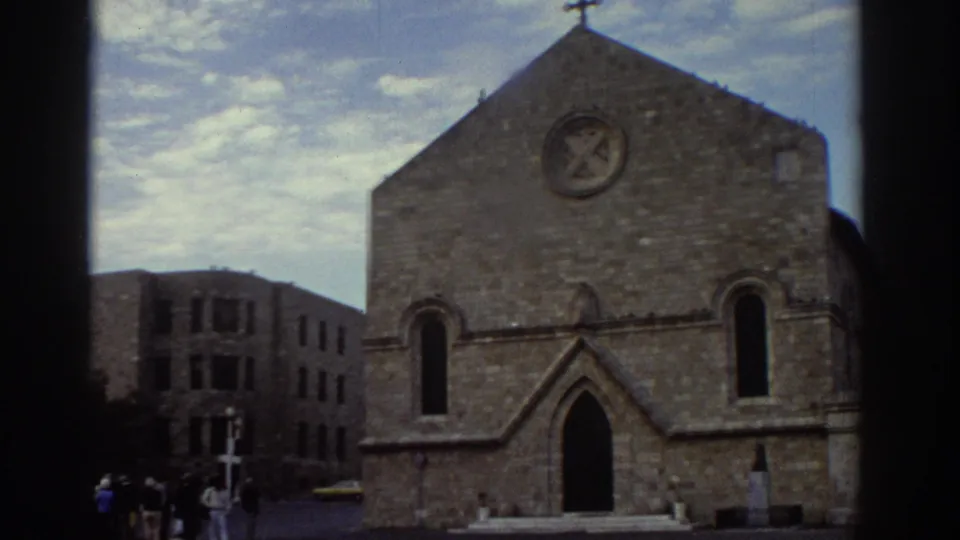 8mm frame from 1976 Greece showing the medieval stone church with rose window and cross, an adjacent stone building, and a figure in white clothing near the entrance at lower right.