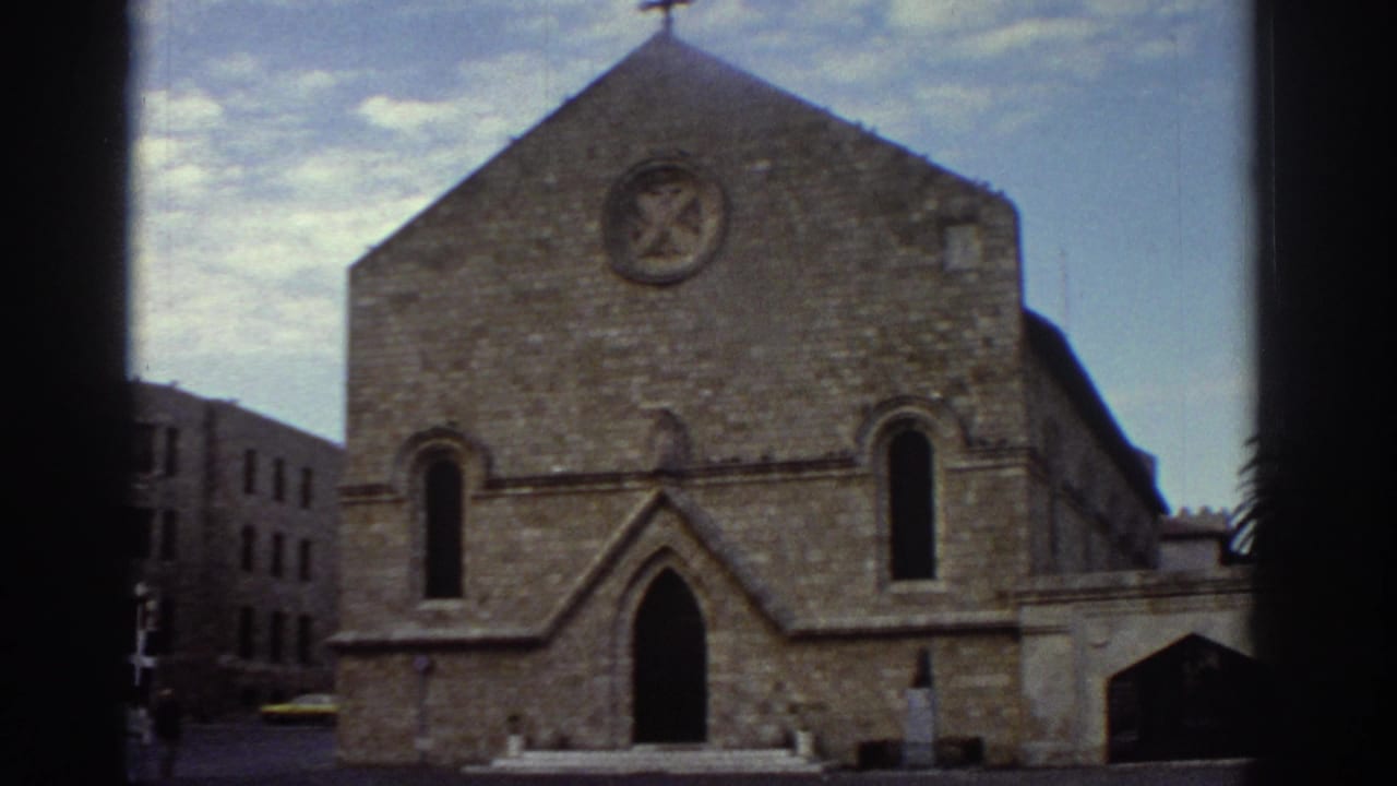 Gothic stone church facade with pointed arched doorways and circular emblem on gable, palm tree at right, likely Greece, c. 1976, 8mm film.