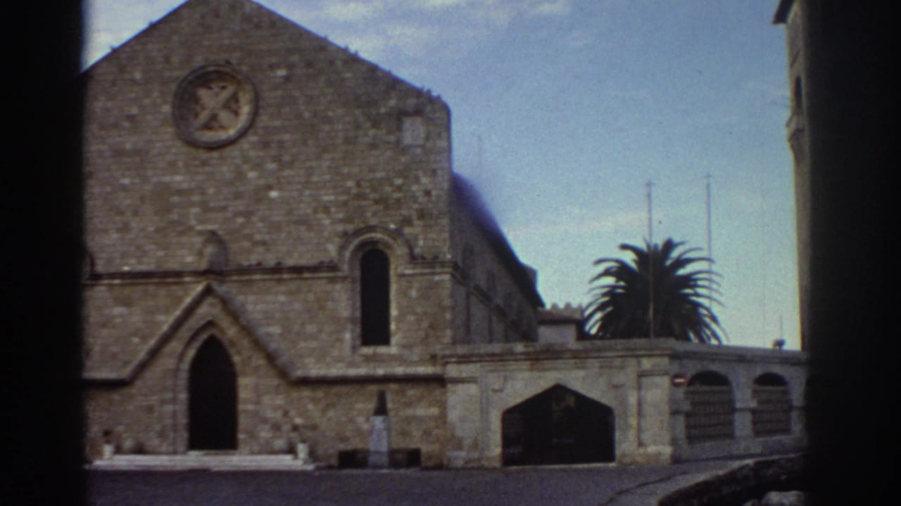 Stone church reframed slightly, Gothic arched doorways and circular emblem visible, palm tree at right, adjacent building at far right edge.