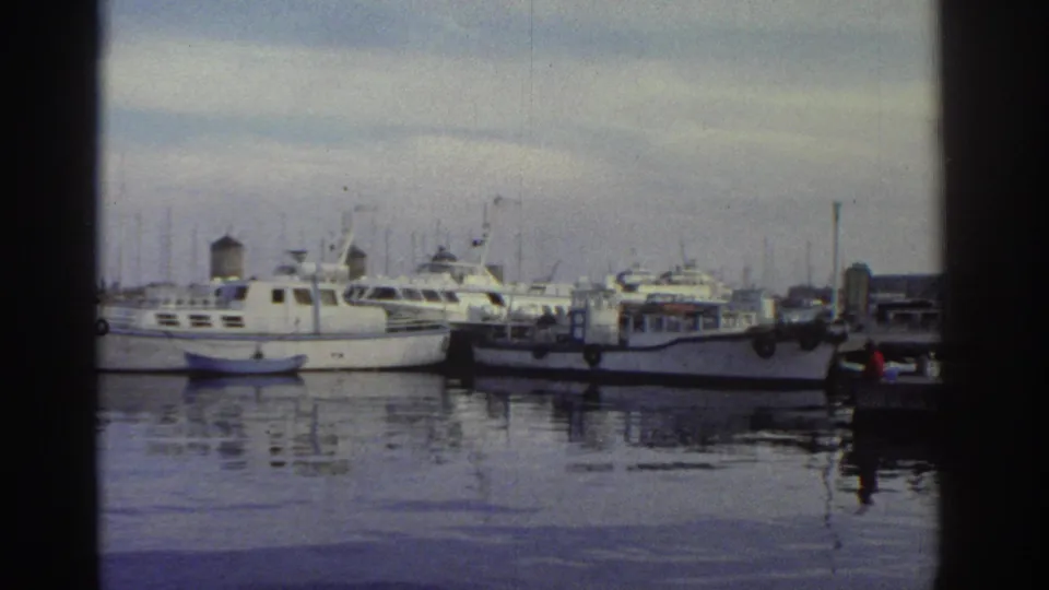 White motor cruisers and yachts moored along a marina quay, calm reflective water, masts, overcast sky, likely Greece, c. 1976, 8mm film.