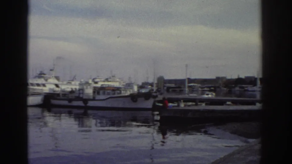 White motor cruisers and yachts moored in a marina under overcast sky, calm water with reflections, likely Greece, c. 1976, 8mm film.