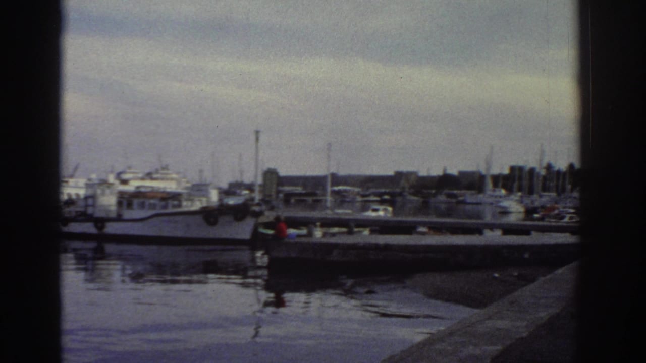 8mm home movie frame of a Greek marina, 1976 — stone pier, densely moored boats, distant harbour buildings under an overcast sky.