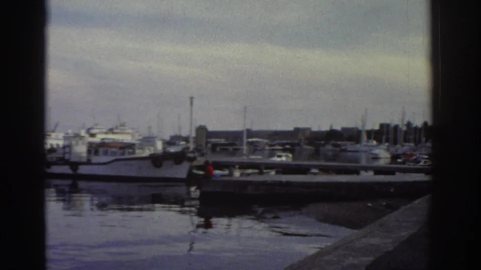 8mm home movie frame of a Greek marina, 1976 — stone pier, densely moored boats, distant harbour buildings under an overcast sky.
