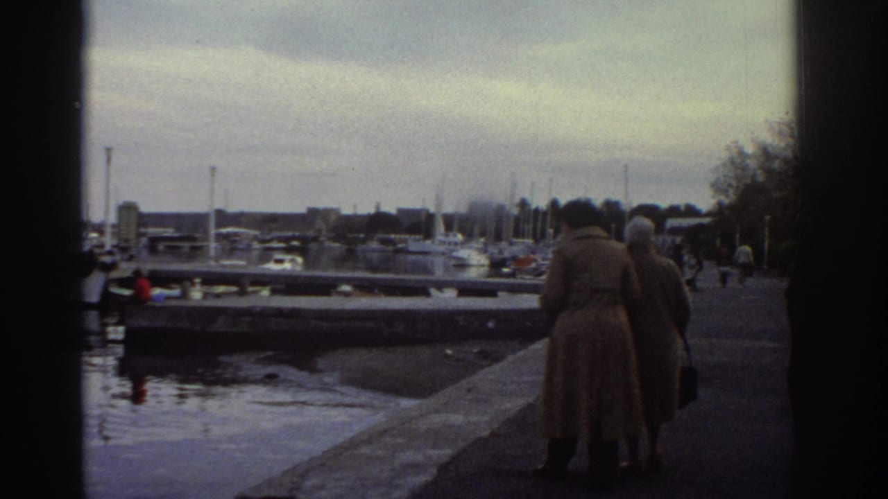 Two figures in long coats walking away from camera along a marina promenade, moored boats in the background.