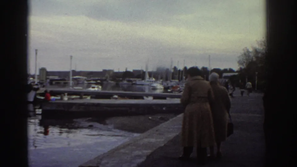 1976 Greece — two women in long coats seen from behind strolling a marina quayside, moored boats and trees visible beyond.