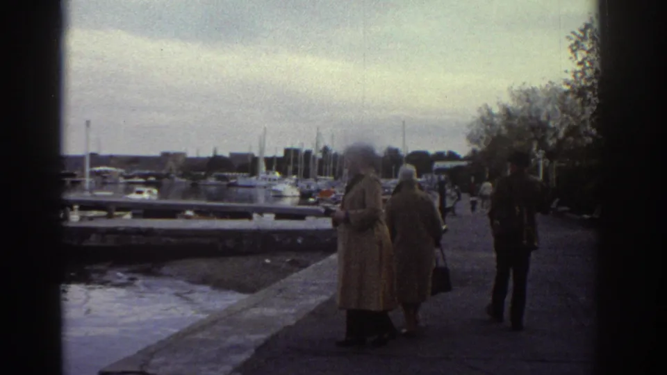 Mid-1970s Greece — closer view of two women in period long coats on a harbour promenade, marina to the left, tree line to the right.